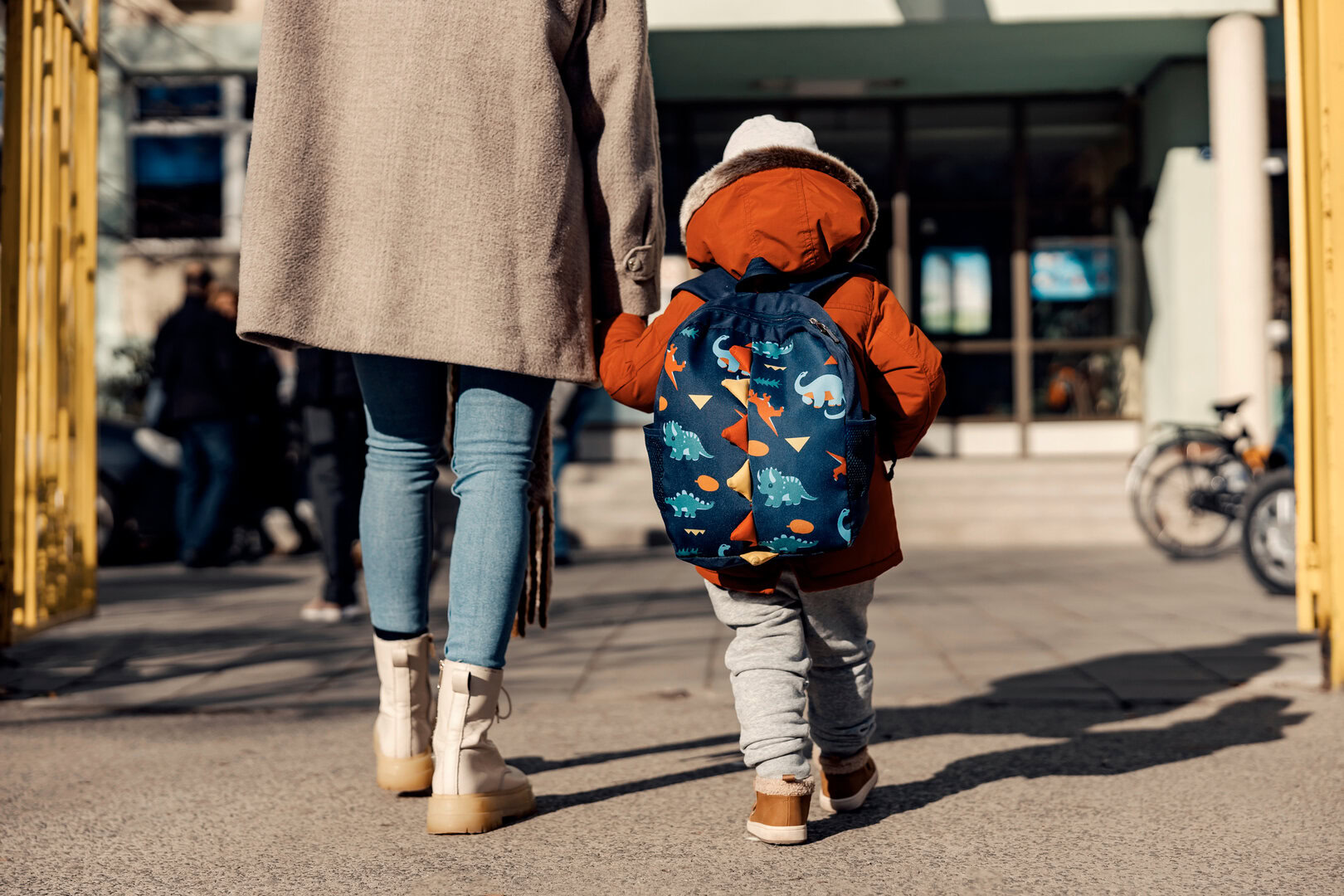 Enfant avec sac à dos accompagné d’un parent se dirigeant vers l’école, illustrant la préparation à la maternelle
